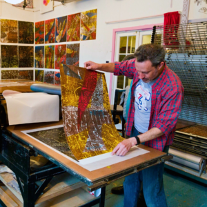 Man holds up a colorful print from the press in a workshop.