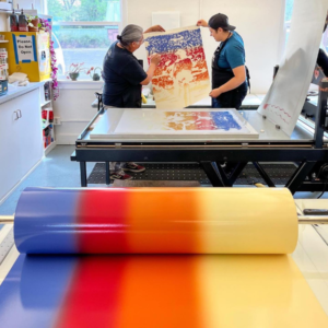A bright roller filled with blue, red, orange, and yellow ink sits in the forefront. Two artists hold up the finished print behind the roller.