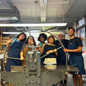 A group of six adults stands together behind a print roller in a print shop. They are all facing the camera and smiling