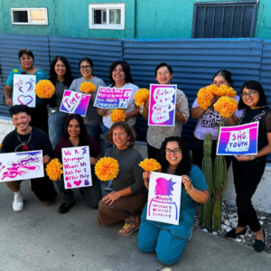 A group of ten or more people stand in two rows, facing the camera, and holding up bright orange paper flowers and colorful prints with political messaging.