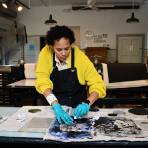 A woman in a bright yellow shirt, a black apron, and blue latex gloves, works at a print making table.